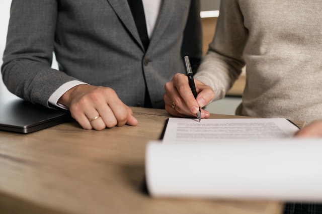 person in a sweater signing a lease contract whole someone else in a suit observes